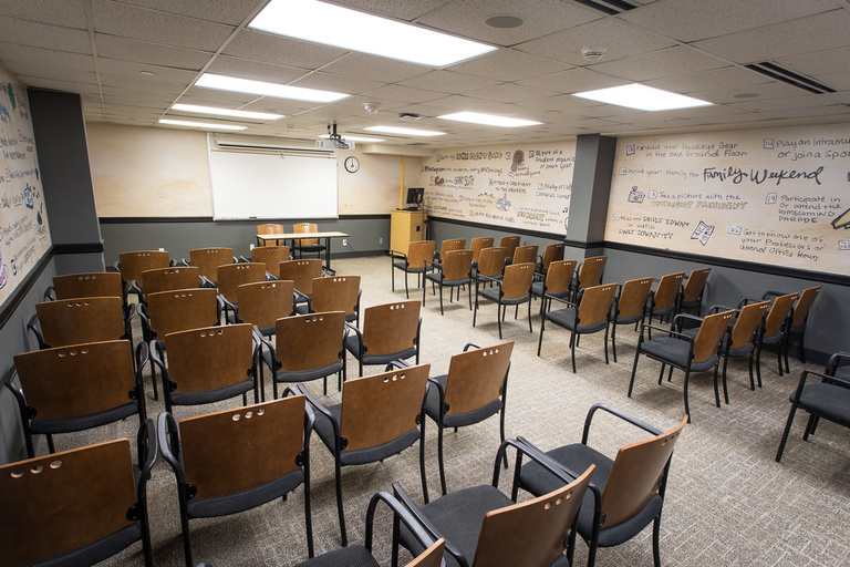 Six Rows of seven to eight chairs facing the front of the room, featuring a table and two chairs, whiteboard, projector, podium and computer.