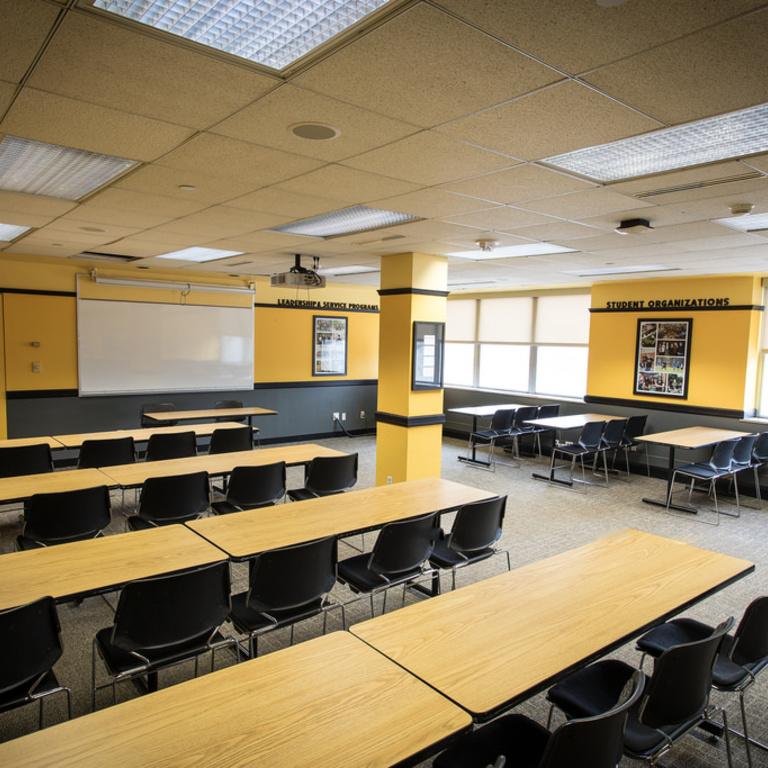 A dozen tables, three seats each, facing front and center table with two seats. Projector and whiteboard.